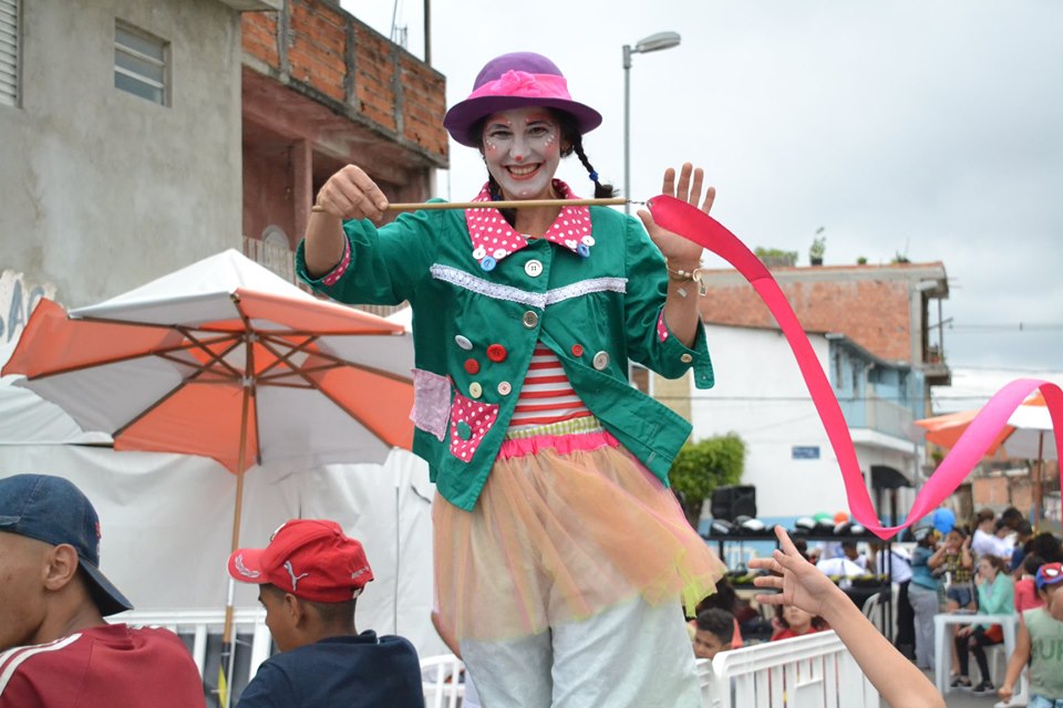 Uma moça, vestida de forma caricata e com rosto pintado, sorri para câmera. Ela está sobre pernas de pau e balança uma fita de cetim Pink. 